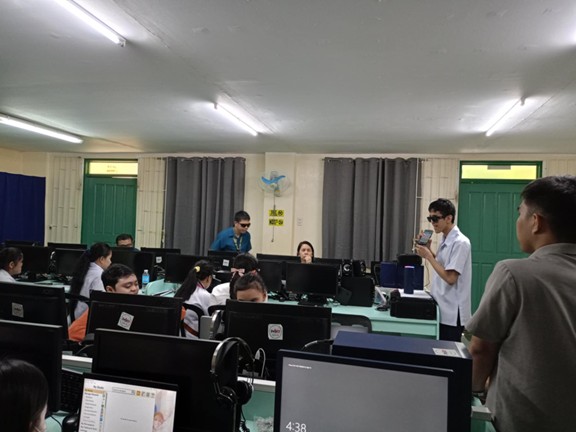 Inside a computer laboratory, several students sit at desktop computers arranged in rows, while teachers and facilitators stand around assisting them. One student wearing sunglasses stands at the front holding a microphone and a smartphone as he speaks to the class. Another teacher, also wearing sunglasses, walks behind the students to provide support. The room has green doors, cream-colored walls, and overhead fluorescent lights, creating a typical classroom setting focused on digital learning.