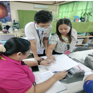hree teachers collaborate around a table during a Braille training session. One teacher, seated and wearing a pink shirt, operates a Braille writer while writing notes in a notebook. Two other teachers stand beside her, leaning in to guide her hand over a sheet of Braille paper. The classroom background includes green curtains, educational materials, and various learning tools, suggesting an active instructional environment.