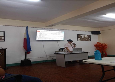 Alt Text: A teacher stands at a podium-style table giving a presentation in a conference room. He is wearing sunglasses and speaking into a microphone while looking at a laptop. A large screen mounted on the wall behind him displays a slide with text. The Philippine flag stands to the left of the screen, and orange decorative plants are on the right.