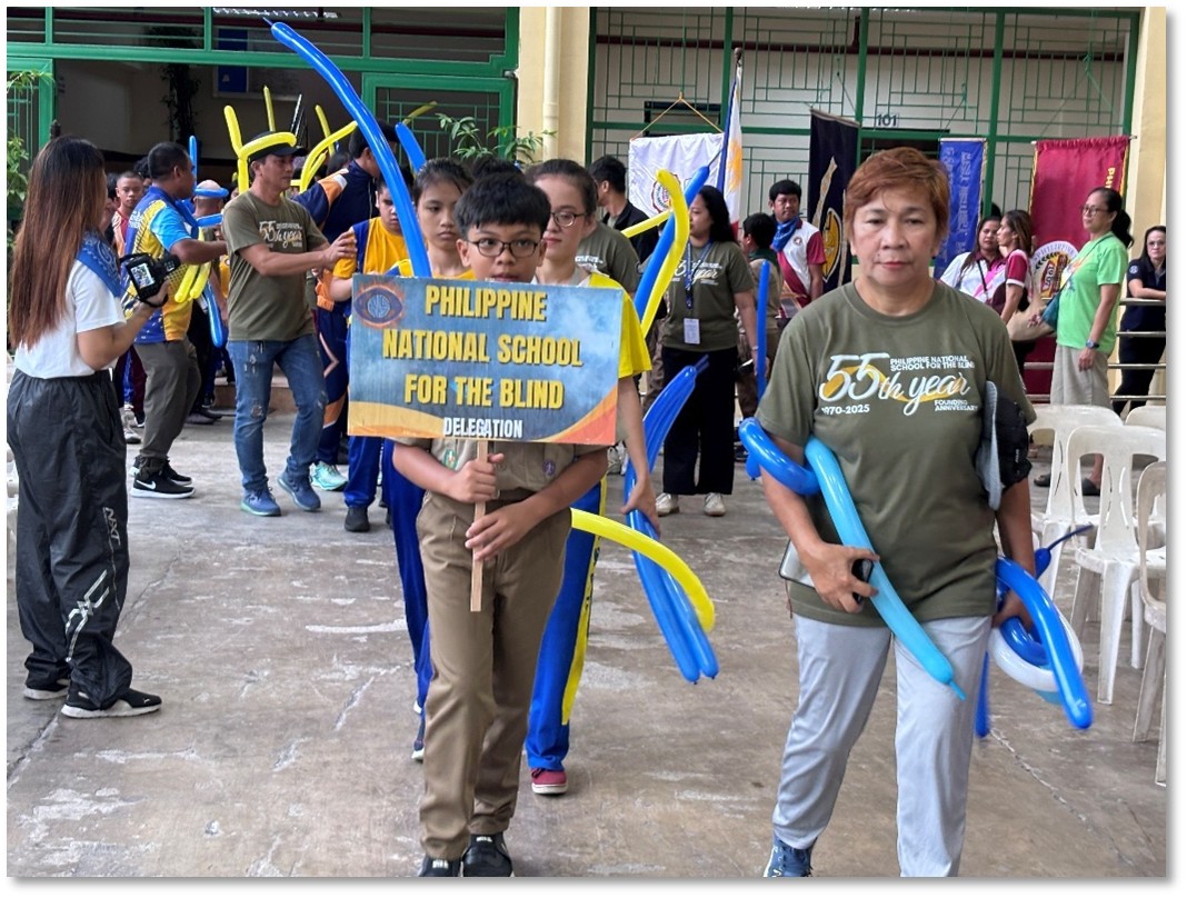 A group of students and teachers from the Philippine National School for the Blind participate in a parade indoors. At the front, a young boy wearing glasses holds a sign that reads “Philippine National School for the Blind – Delegation.” Behind him, other students walk in line while holding long blue and yellow balloon decorations. Teachers and staff wearing olive-green shirts with “55th Year” printed on them accompany the group. More participants and school banners can be seen in the background as the delegation enters the venue.