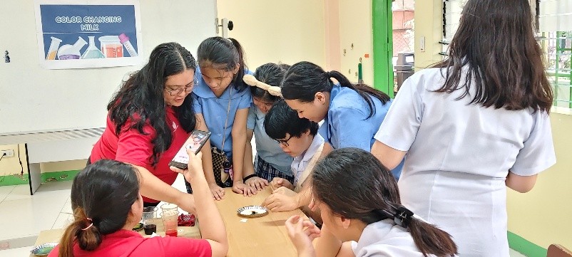 A group of students gather around a table as a teacher guides them through a hands-on science activity. The students lean in closely, touching or observing materials placed on a small plate at the center. One student records the experiment using a smartphone. A poster labeled “Color Changing Mix” is displayed on the board in the background, indicating the activity theme. The classroom is bright, with cream-colored walls and green doors and windows.