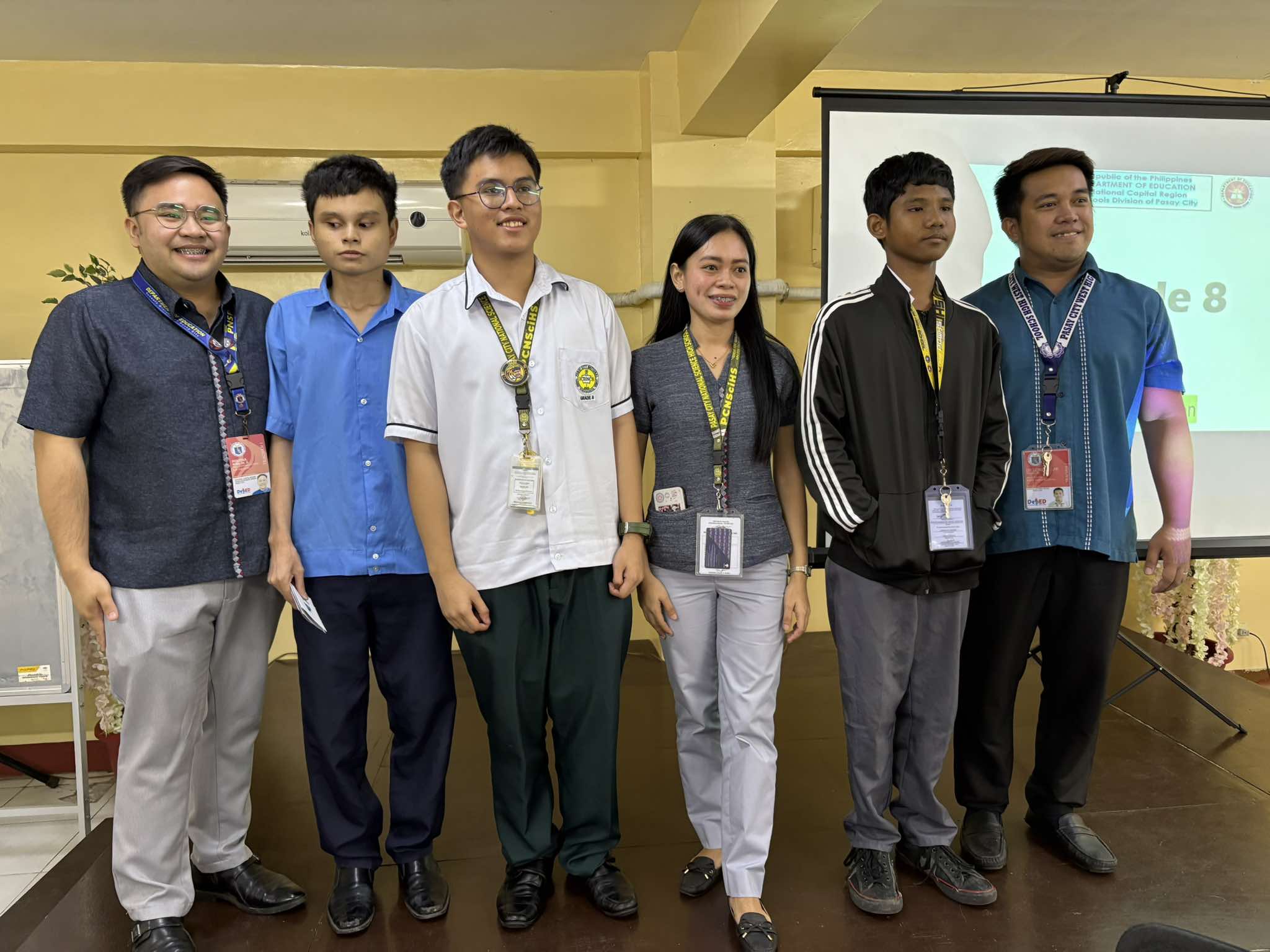Three students stand in front of a classroom stage with three teachers behind them, all smiling for a group photo. The boy on the left wears a blue uniform shirt and dark pants, with a face mask hanging from his neck. The girl in the middle wears a white blouse and a red checkered skirt with an ID lanyard. The boy on the right wears a white polo shirt, brown pants, eyeglasses, and a blue lanyard. Behind them, the three teachers are standing close and smiling. On the right side of the image is a projector screen displaying a partially visible slide.