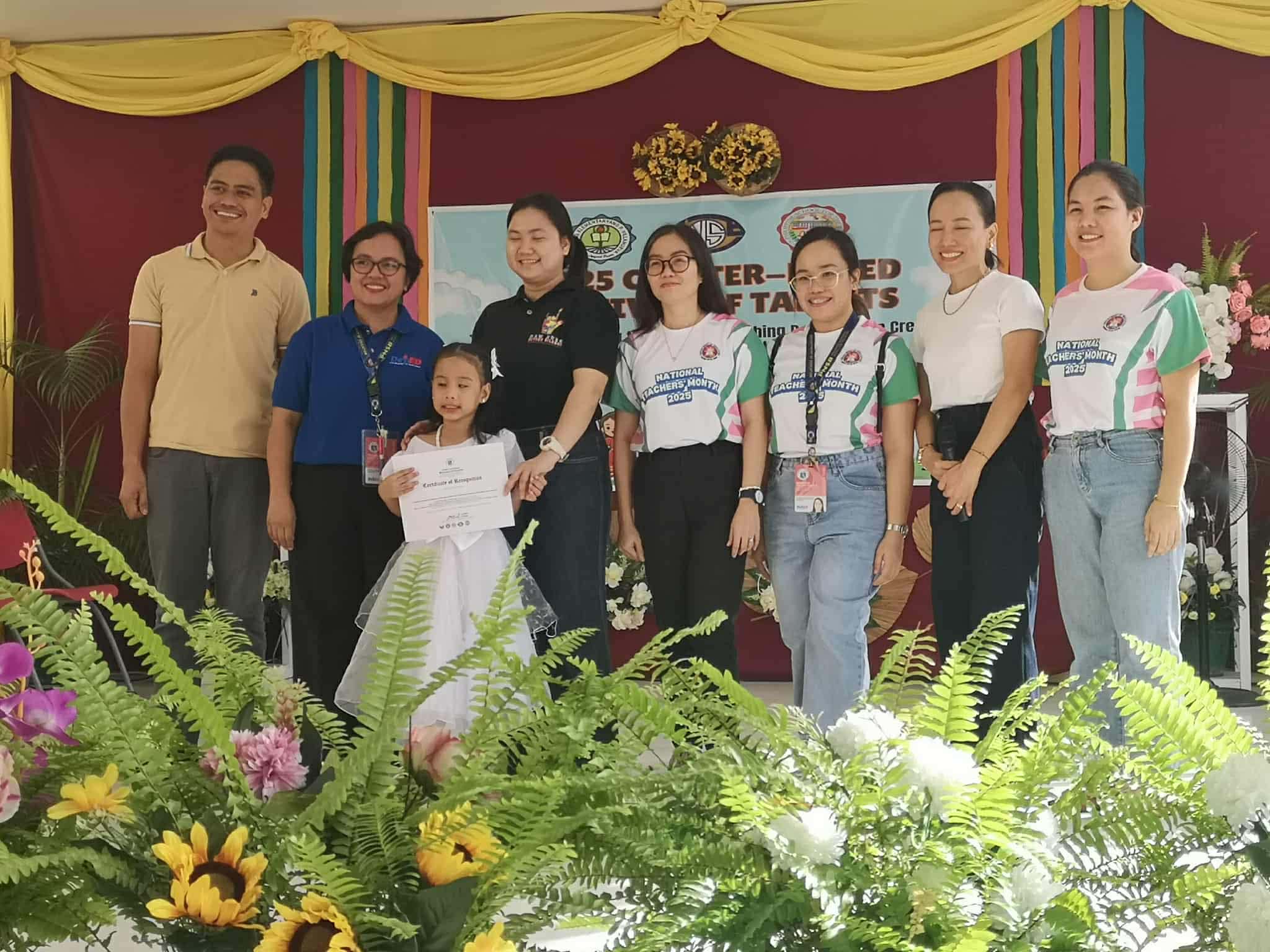 A young girl in a white dress stands at the center of a stage holding a certificate, smiling as she is recognized during an awarding ceremony. Behind her is a row of eight adults—teachers and staff—standing and smiling proudly. Some wear National Teachers' Month 2023 event shirts, while others wear casual or semi-formal attire.
